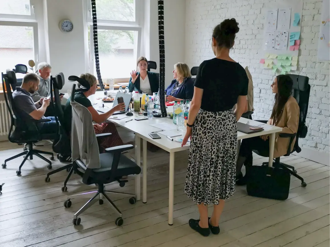 A group of people sitting at a table during a discovery workshop in a bright modern office. There are matrices with drawings and sticky notes hanging on the wall. Snacks and workshop materials on the table